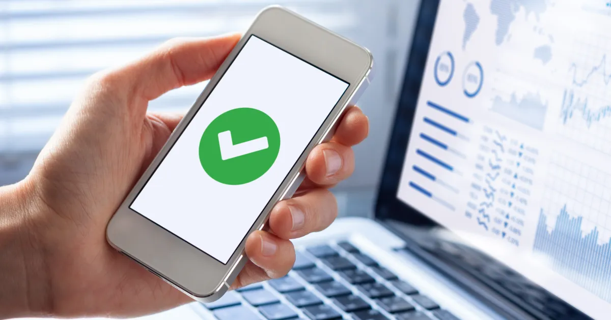 A phone showing a checkmark in front of a computer with supply chain analytics.